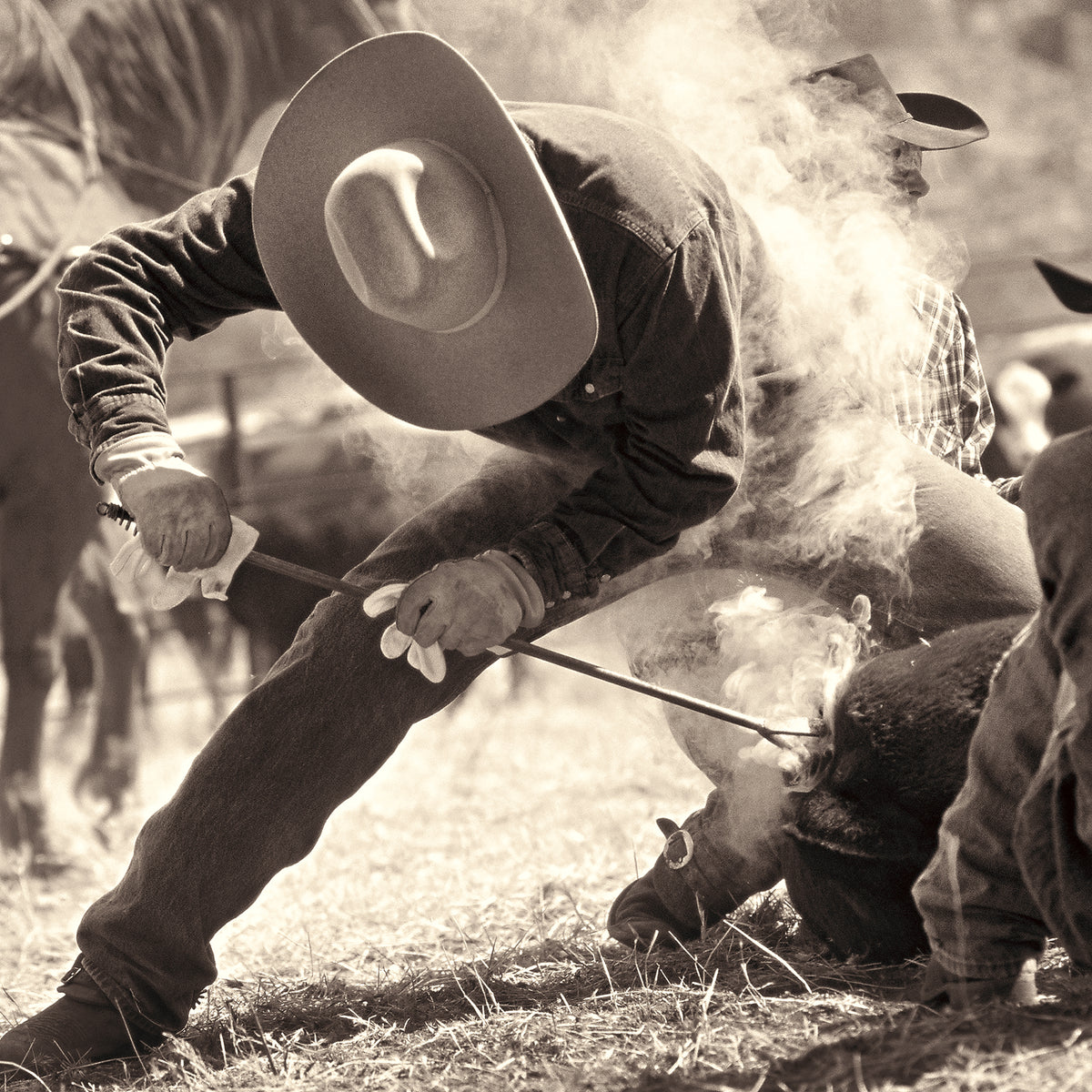 A sepia-toned action scene showing a cowboy in a wide-brimmed hat leaning forward and using a long-handled tool over a smoking animal in an outdoor ranch setting.